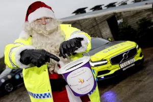 PC Claus, The Santa in Police uniform showing a charity bucket, with a police car behind.